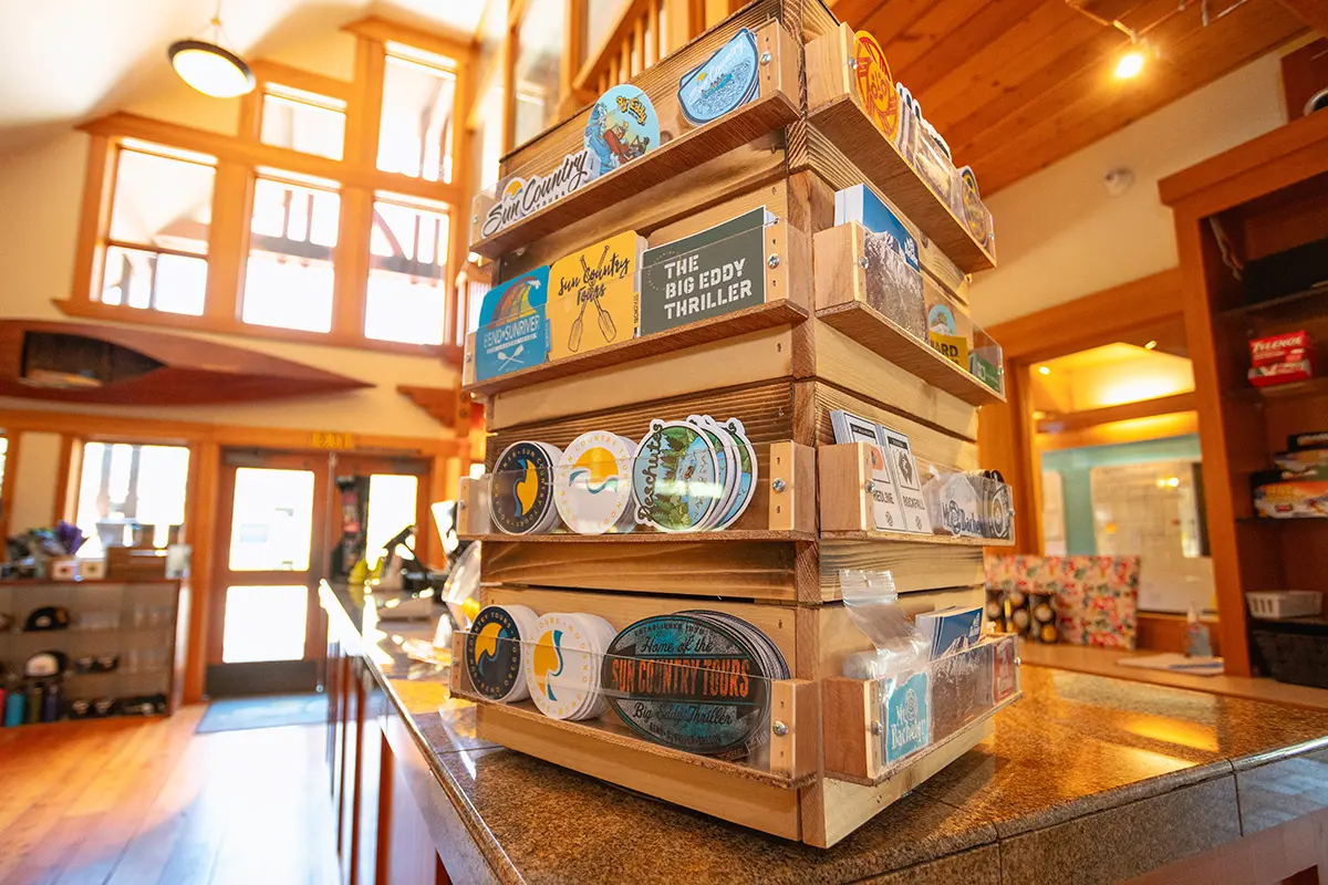 Display of colorful stickers and postcards at the Sun Country Tours retail store in Bend, Oregon. The wooden display rack features various designs highlighting the outdoor adventures and scenic locations associated with Sun Country Tours. The store's interior includes large windows, natural lighting, and wooden accents, creating a welcoming and rustic shopping environment for visitors seeking souvenirs and gear for their outdoor experiences.