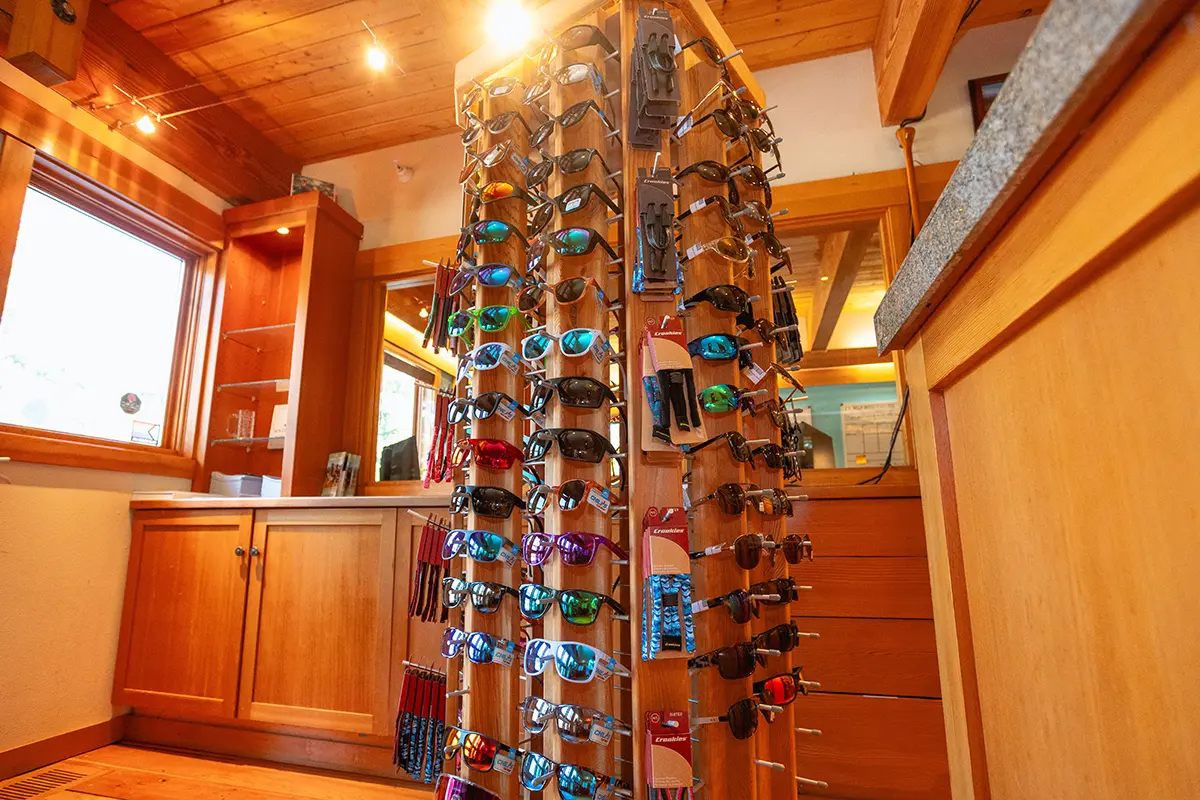 Display of various sunglasses at the Sun Country Tours retail store in Bend, Oregon. The wooden stand features a wide selection of sunglasses with different styles and lens colors, catering to outdoor enthusiasts. The store's interior includes natural wood finishes, ample lighting, and large windows that create a bright and inviting shopping atmosphere for visitors looking to gear up for their outdoor adventures.
