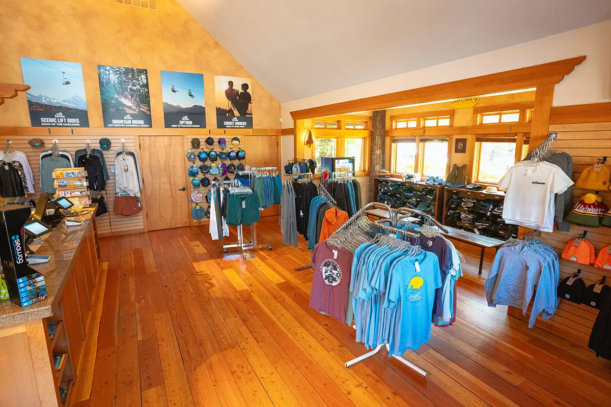 Wide view of the Sun Country Tours retail store in Bend, Oregon, showcasing a variety of apparel and gear on display. The store features wooden floors and walls, creating a warm and inviting atmosphere. The racks are filled with branded t-shirts, hats, and hoodies, while promotional posters highlight the various outdoor activities offered by Sun Country Tours. The well-organized space is designed to provide an excellent shopping experience for visitors looking to gear up for their adventures.