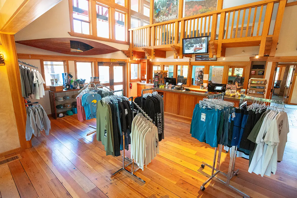 Interior view of the Sun Country Tours retail location in Bend, Oregon, showcasing a bright and welcoming shop with wooden floors, racks of branded apparel, and various merchandise. The store features large windows providing natural light, wooden accents, and a cozy, inviting atmosphere perfect for outdoor enthusiasts looking to gear up for their adventures.